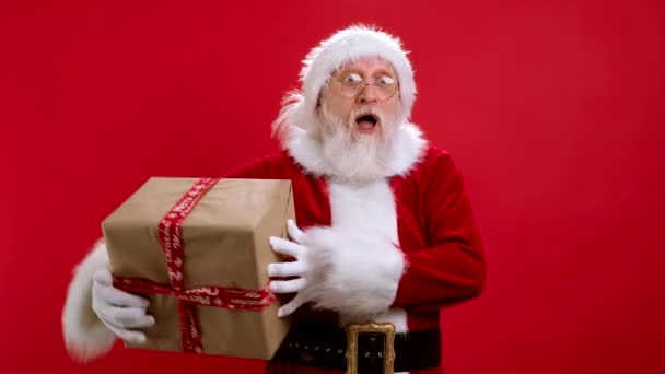 Babbo Natale divertente in costume di Natale con scatole regalo su sfondo rosso guarda fotocamera e cattura scatole di regali. Babbo Natale allegro lancia regali. Buon Natale e regali da San Nicola. — Video Stock