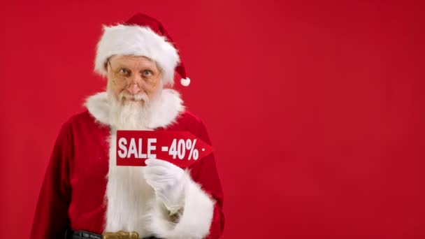 Kerstman Houdt Teken met inscriptie Sale -40 uit, wijst zijn vinger op een lege ruimte Mock up en kijkt naar korting in de camera en glimlacht op de rode achtergrond. Grote korting, kerstvakantie verkoop. — Stockvideo