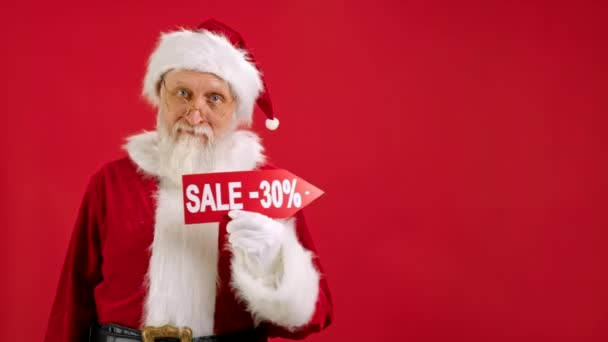 Święty Mikołaj trzyma znak z napisem sprzedaż -30 off, wskazuje palcem na puste Kosmiczna makieta i patrzy na Rabat w aparacie i uśmiecha się na czerwonym tle. Duże rabaty, świąteczne sprzedaży. — Wideo stockowe