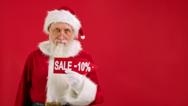 Der Weihnachtsmann hält ein Schild mit der Aufschrift Sale -10 off, zeigt mit dem Finger auf eine Leerraum-Attrappe und blickt in die Kamera und lächelt auf rotem Hintergrund. Großer Rabatt, Weihnachtsgeschäft. — Stockvideo