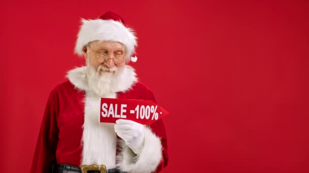 Kerstman houdt ondertekenen met inscriptie Sale -100 uit, wijst zijn vinger op een lege ruimte Mock up en kijkt naar korting in de camera en glimlacht op de rode achtergrond. Grote korting, kerstvakantie verkoop. — Stockvideo