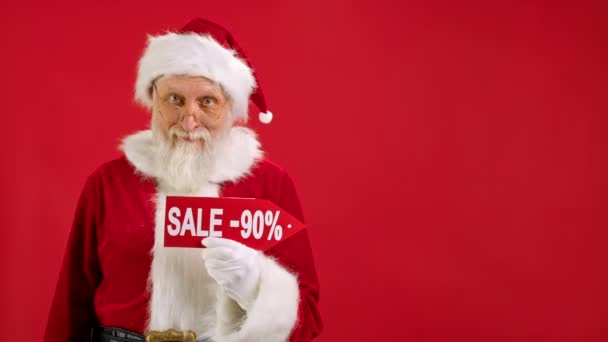 Santa Claus sostiene el cartel con la venta de inscripción 90 de descuento, señala su dedo en un espacio vacío se burlan y mira el descuento en la cámara y sonríe en el fondo rojo. Descuento grande, Ventas de vacaciones de Navidad. — Vídeos de Stock