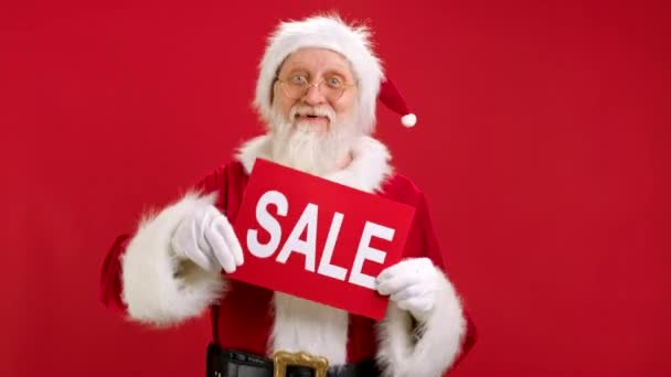 クリスマスセールサンタクロースが赤い背景にバナーを持っている彼の手で販売を手紙とカメラを見て。サンタクロースからメリークリスマスセール、バナーモックアップと踊る販売サインと笑顔. — ストック動画