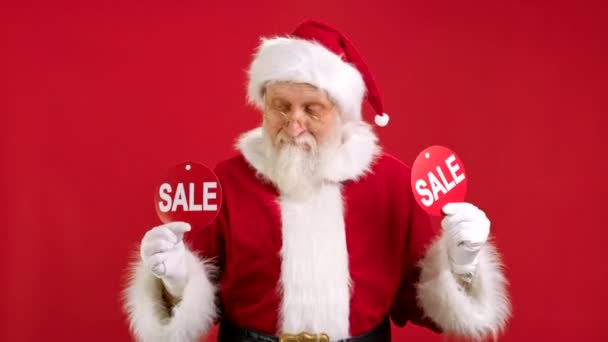Weihnachtsverkauf. Der fröhliche Weihnachtsmann tanzt und freut sich über den Weihnachtsverkauf und hält zwei Banner mit der Aufschrift SALE in die Kamera auf rotem Hintergrund. — Stockvideo