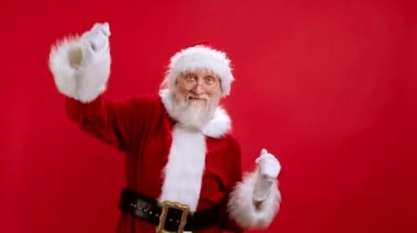 Noel Baba kostümlü beyaz adam dans gösterisinde eğleniyor. Yeni yıl ve Noel tatili kavramı. Neşeli Mutlu Noel Baba Stüdyo Kırmızı Arkaplanda dans ediyor, Neşeyle ellerini sallıyor.