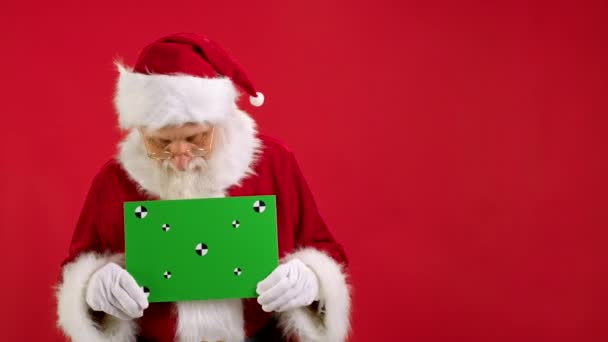 快乐的圣诞老人拿着绿色屏幕横幅追踪点寻找复制空间。白纸绿色屏风用他的手和地方在红色背景上的文字或广告挥手致意. — 图库视频影像