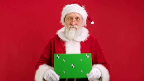 Fröhlich wirbt der Weihnachtsmann mit einem Banner, hebt eine grüne Attrappe mit Fahndungspunkten in die Höhe, blickt auf ein grünes Banner und schwenkt seinen Kopf. Layout-Werbung mit Green Screen für Ihren Text. — Stockvideo
