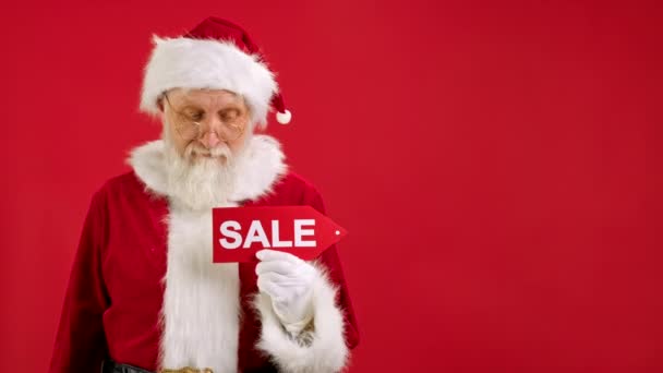 Der Weihnachtsmann hält ein Schild mit der Aufschrift Verkauf, zeigt mit seiner zustimmenden Handgeste auf eine Leerraum-Attrappe und blickt in die Kamera und lächelt auf rotem Hintergrund. Großer Rabatt, Weihnachtsgeschäft. — Stockvideo