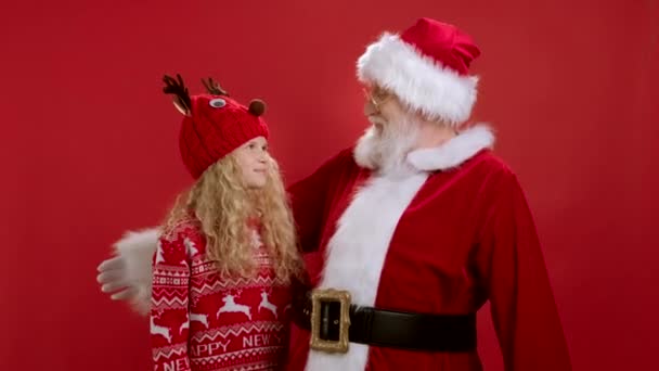 Симпатичный Санта-Клаус в красной одежде обнимает маленькую блондинку в красной шапочке и свитере, улыбается и машет руками на красном фоне. Объятия и дружба Санты и ребенка во время рождественских праздников. — стоковое видео