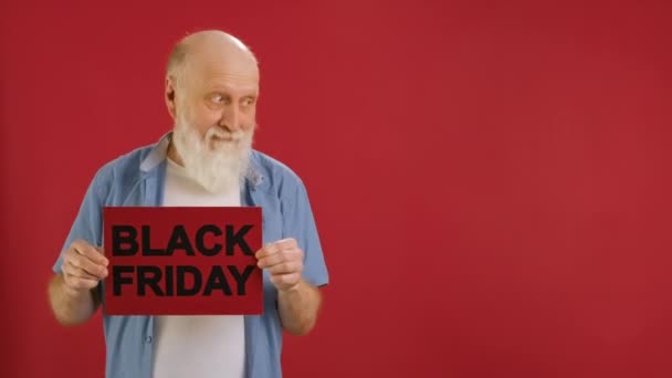 Vidám idős nagyapa, kopasz fej és szakáll fehér póló és kék ing emeli banner felirattal BLACK FridAY örül a nagy eladó vagy kedvezmény a boltban, és tánc piros háttér. — Stock videók