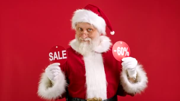 Boże Narodzenie SPRZEDAŻ -60. Wesoły Święty Mikołaj tańczy i raduje się ze świątecznej wyprzedaży, trzymając dwa transparenty z napisem SPRZEDAŻ i -60 Off Pokazując napisy do kamery na czerwonym tle. — Wideo stockowe