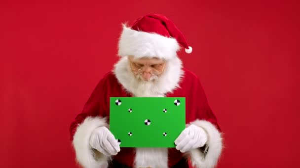 Santa Claus Shows an Approving Thumbs-up Gesture With Space for Text or Advertising on Red. Cheerful Santa Claus Holding Green Screen Banner Tracking Points for Copy Space. Blank Green Screen Board. — Stock Video