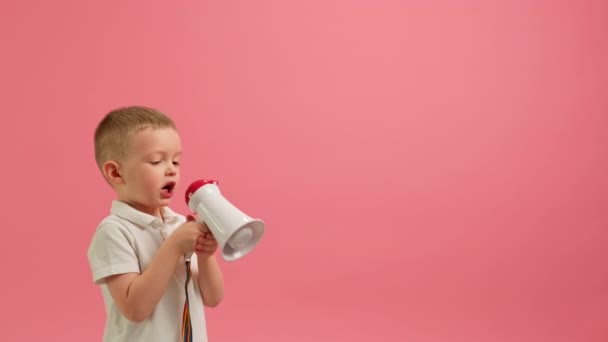 Kleine blonde blanke jongen in wit T-shirt schreeuwt luid in witte megafoon, plaats voor tekst of reclame op roze achtergrond. Kind schreeuwt in luidspreker, informeert over grote verkoop en kortingen. — Stockvideo