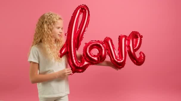 Young caucasian blonde girl holds in her hands a red inflatable balloon with inscription LOVE smiles and happily dances celebrating Valentines Day. Concept of congratulating Valentines Day. — Vídeo de Stock