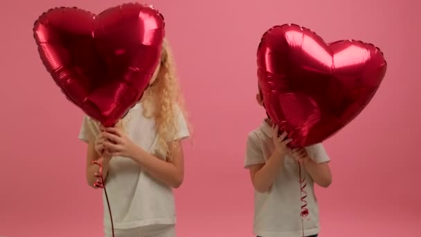 Two cute blonde children boy and girl are holding in their hands red balloons in shape of heart have fun on Valentines Day or Mothers Day and look out from behind helium balloons on pink background. Video Clip