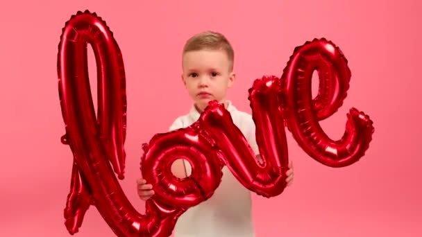 Young caucasian blonde boy holds in her hands red inflatable helium balloon with inscription LOVE smiles and happily dances celebrating Valentines Day. Concept of congratulating loving day. — Vídeo de Stock