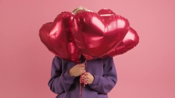 Portret van vrolijke glimlachende vrouw die geniet van Moederdag of Valentijnsdag verbergt haar gezicht met rode gel ballen in de vorm van hart op roze achtergrond, gluurt uit rode harten. liefde concept, gelukkig gezin. — Stockvideo