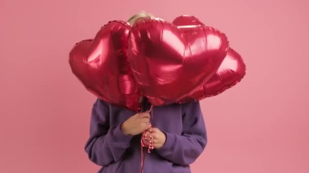 Ritratto di allegra donna sorridente che si gode la festa della mamma o San Valentino nasconde il viso con palline di gel rosso a forma di cuore su sfondo rosa, sbircia fuori dai cuori rossi. concetto di amore, famiglia felice. — Video Stock