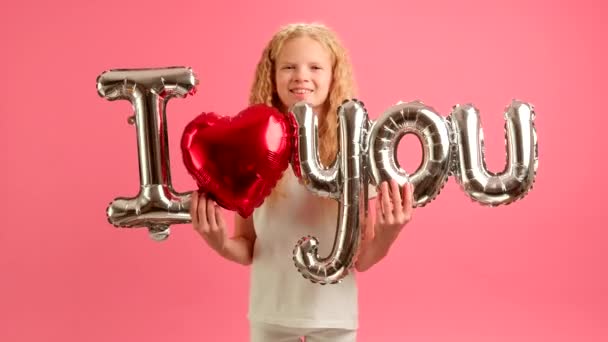 Young caucasian blonde girl holds in her hands red and silver inflatable balloon with inscription I LOVE YOU smiles and happily dances celebrating Valentines Day. Concept of congratulating loving day. — стоковое видео