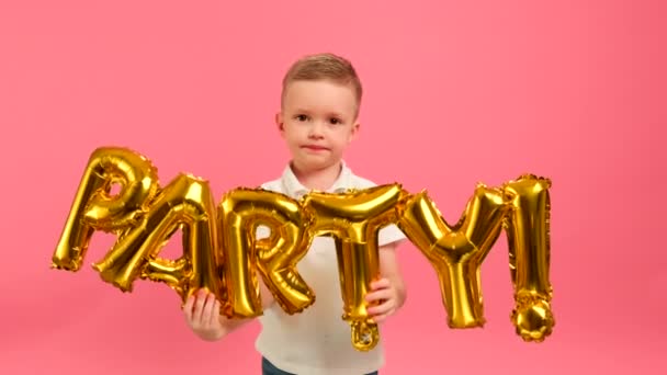 Glad blond vit pojke hoppar dans jublar på fest hålla i sina händer gyllene brev med inskription PARTY på rosa bakgrund. Pojke visar inskription PARTY i guld ballong bokstäver. — Stockvideo