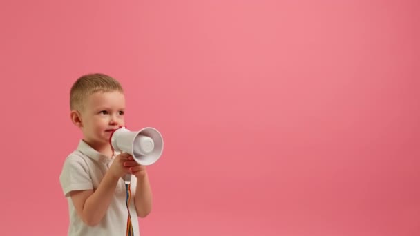 Boy agitates by shouting into loudspeaker and pointing his finger to side on pink background. Blond boy in white T-shirt shouts into megaphone and points to an empty advertising space with his hand. — Vídeo de Stock