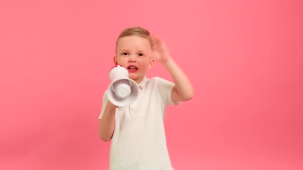Kid shouts emotionally into loudspeaker waving his hand. Portrait 5 years boy in white T-shirt shouts expresses his dissatisfaction, swears masher menacingly shouts into megaphone on pink background. — Stockvideo