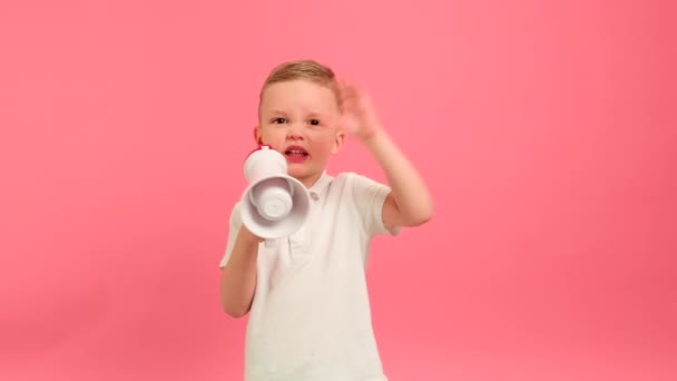 Kid shouts emotionally into loudspeaker waving his hand. Portrait 5 years boy in white T-shirt shouts expresses his dissatisfaction, swears masher menacingly shouts into megaphone on pink background. — стоковое видео