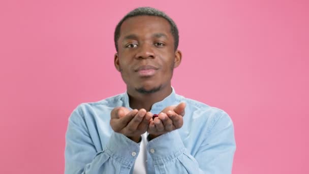Retrato de homem adolescente alegre afro-americano despreocupado em camisa azul soprando confete em forma de coração vermelho, desfrutando de aniversário ou dia dos namorados, humor festivo. Estúdio interior filmado em fundo rosa. — Vídeo de Stock