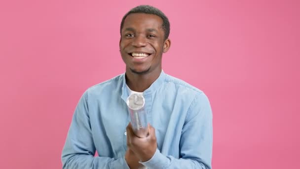 Happy 20 year African American teenager, man in denim shirt, is smiling, celebrating birthday or winning, explodes firecracker with confetti in to camera, slow motion on pink studio background. — Stock video