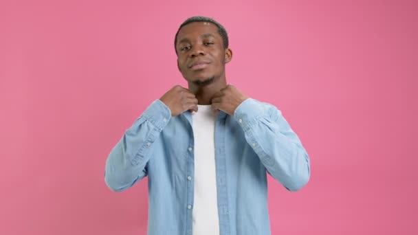 Handsome smiling cute young African American 20 years old in denim shirt and white T-shirt poses and blows air kiss to camera on pink studio background. Emotions of love, cheerful person. — Stok video