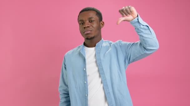 Unsatisfied African American 20 years old in denim shirt, white T-shirt shows thumbs down, dislike, negative feedback, covers his face with his hands shot in studio on pink background. — стоковое видео