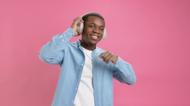 Χαρούμενος Αφρο-Αμερικανός άνδρας με denim πουκάμισο και ακουστικά Bluetooth στο κεφάλι του ακούει μουσική, χαμογελάει με ευχαρίστηση και ραπάρει σε hip-hop ρυθμούς σε ροζ φόντο. Ράπερ πορτραίτου αργής κίνησης. — Αρχείο Βίντεο