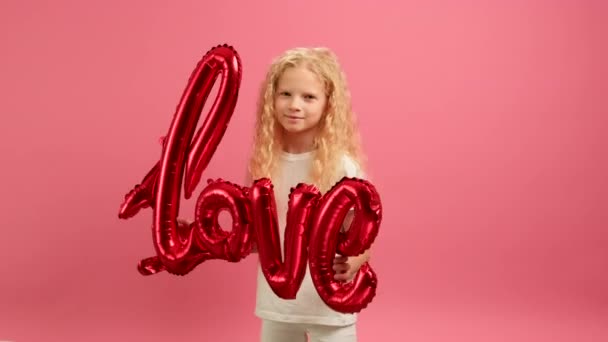 Chica sostiene globo con letras AMOR en sus manos y baila alegremente en el Día de San Valentín. Felicidades por las vacaciones del amor. Chica joven con el pelo largo bailando con una bola de gel de aire sobre fondo rosa. — Vídeos de Stock