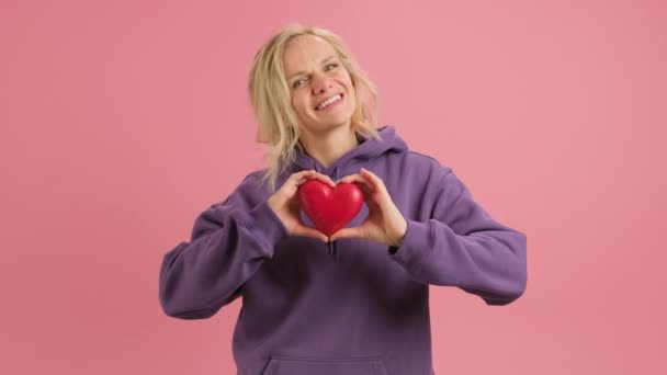 Young woman holds red paper heart on her chest. Symbol romantic love. Woman in purple hoodie with red heart in her hands on pink background. Concept of falling in love, Valentines Day or Mothers Day. — 图库视频影像