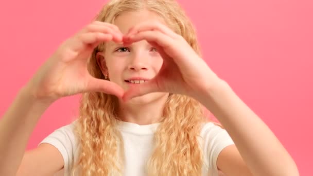 Smiling young girl volunteer showing hands sign heart shape looking at camera on pink background. Healthy heart health life insurance, love and charity, voluntary social work, organ donation concept. — Stock video