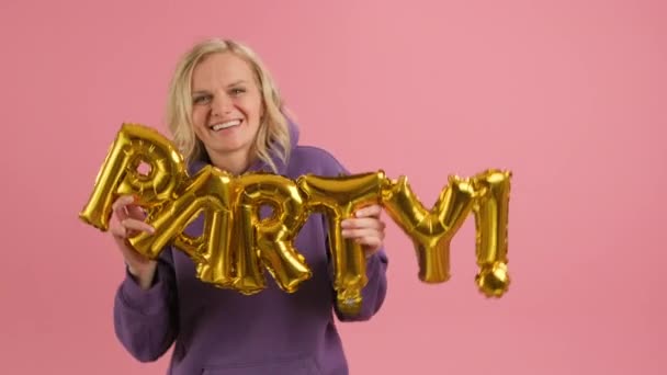 Slow motion shot happy blonde smiling woman in Very Berry hoodie is having fun at party or birthday holding balloon golden inscription party in hands and dancing standing on pink background. — Stockvideo