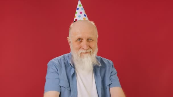 Szczęśliwy 60-letni emerytowany dziadek z gwizdkiem i stożkiem na głowie eksploduje konfetti na czerwonym tle i cieszy się. Staruszek świętuje urodziny wysadzając petardę konfetti w kamerę. — Wideo stockowe