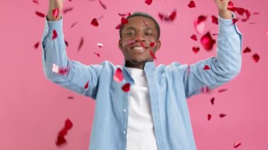 Yavaş çekim gülümseyen Afro-Amerikan Sevgililer Günü 'nde veya Anneler Günü' nde herkesi tebrik eder, kırmızı kalplerden konfeti atar, parmaklarıyla kalp hareketi yapar, pembe arka planda aşkı gösterir..