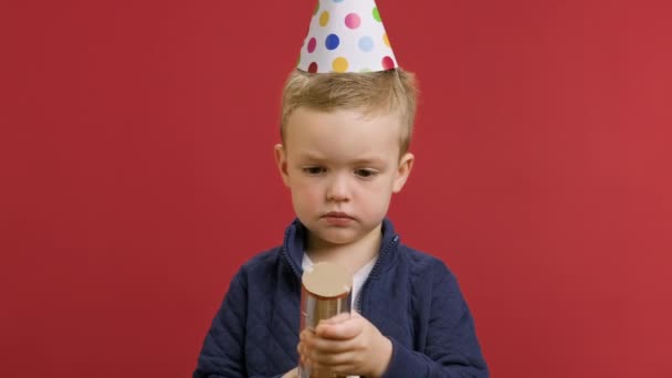 Happy little birthday boy met feestelijke kegel op zijn hoofd viert zijn verjaardag explodeert rotje met confetti en glimlacht geniet van feestelijke dag op rode studio achtergrond. Gelukkige verjaardag.. — Stockvideo