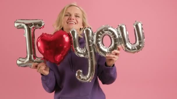 Mujer rubia caucásica feliz sostiene en sus manos globo inflable rojo y plata con inscripción I LOVE YOU sonríe y baila felizmente celebrando el Día de San Valentín o el Día de las Madres. — Vídeos de Stock