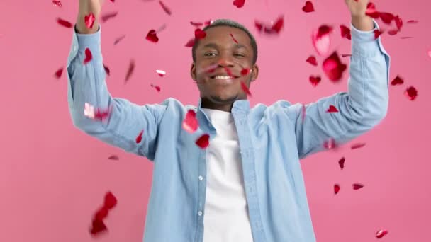 Slow motion sonriendo afroamericano felicita a todos en el Día de San Valentín o el Día de las Madres, vomita confeti de corazones rojos, haciendo gesto del corazón con los dedos, mostrando amor, en fondo rosa. — Vídeos de Stock