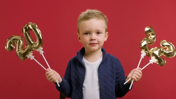 Portrait de garçon réjouissant 2022 année tigre entrant debout sur fond de studio rouge. mignon blond caucasien enfant garçon tient or nombres gonflables 2022 dans ses mains et couvre son visage avec des chiffres. — Video