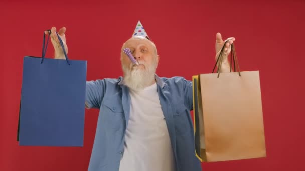 Boldog, idős, szakállas férfi vidáman táncol, csomagolóanyagokat tart a kezében, hogy eladhassa őket. Nagyapa boldog vásárlás, hencegő papírzacskók, fújás papír síp piros háttér. — Stock videók