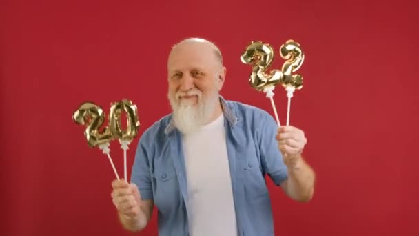 Portrait of cheerful smiling bearded grandfather holding in his hands golden numbers 2022 of new year tiger smiling cheerfully waving inflatable figures dancing on red studio background. — Stock Video