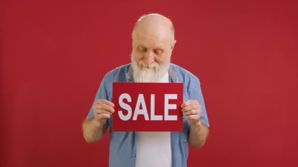 Portrait Elderly cheerful grandfather is dancing with Sale banner in hands on red studio background. Big sale on Black Friday, big discount, Christmas sale. — Stock Video