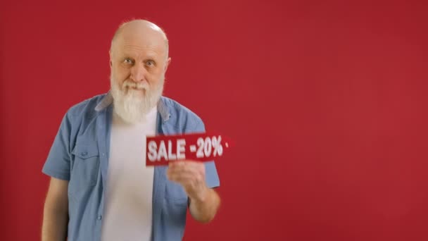Old bearded man Holds Sign With Inscription Sale -20 off, Points His Finger at Empty Space Mock up and Looks at Discount in Camera and Smiles on Red Background. Μεγάλες εκπτώσεις τις μαύρες Παρασκευές. — Αρχείο Βίντεο