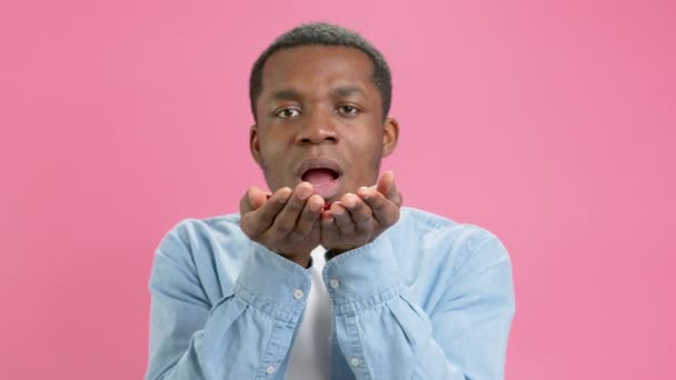 Portrait of carefree African American cheerful teenage man in blue shirt blowing red heart shaped confetti, enjoying birthday or valentines day, festive mood. Indoor studio shot on pink background. — Stockvideo