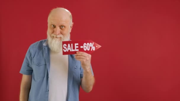 Old bearded man Holds Sign With Inscription Sale -60 off, Points His Finger at Empty Space Mock up and Looks at Discount in Camera and Smiles on Red Background. Μεγάλες εκπτώσεις τις μαύρες Παρασκευές. — Αρχείο Βίντεο