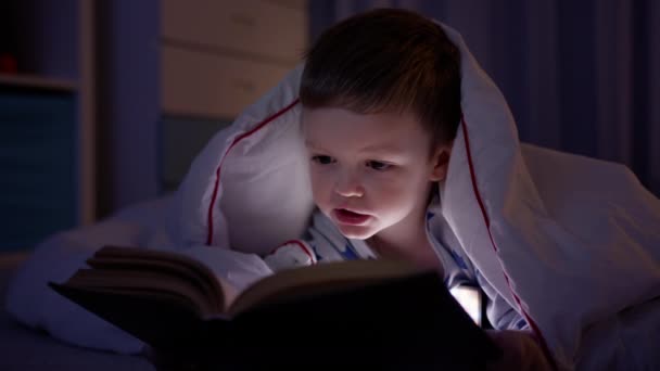 Portrait small blond boy child in pajamas and covered with white blanket lies on floor of dark night room and reads bedtime story. Kid learns lessons, reads book, engaged in self-development. — Stock Video