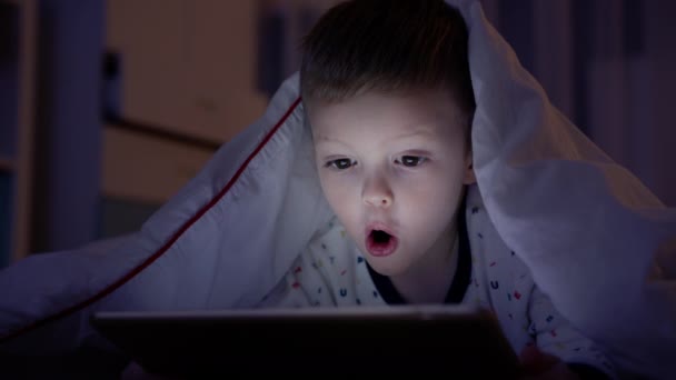 小さな金髪の少年は感情的な恐怖を経験し、ラップトップで怖い映画を見ながら彼の顔を手で覆い、ベッドの上の夜の家で白い毛布の下で夜に横たわっています。子供は怖い映画. — ストック動画